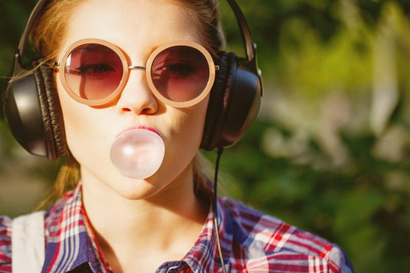 Young hipster girl listening to music on headphones in a summer park. Portrait close-up with chewing gum. Warm toning. The concept of cheerful youth.